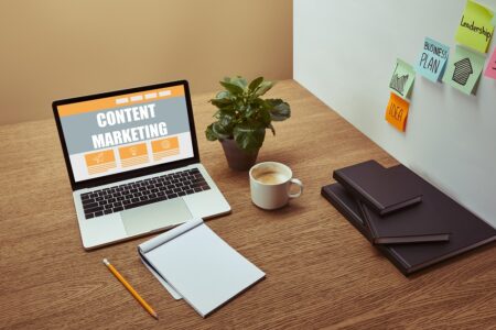 A laptop displaying "Content Marketing" on a wooden desk highlights the power of leveraging content marketing, surrounded by a notebook, pencil, coffee cup, plant, file folders, and sticky notes on the wall in the background.