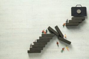 Miniature figures climb uneven stairs toward a briefcase, with some falling from broken steps—symbolizing career challenges. Just as in work, the benefits of using paid ads can help overcome obstacles and reach your goals more efficiently.