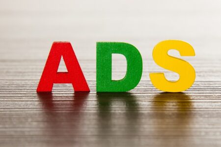 Three wooden letters, A in red, D in green, and S in yellow, spell "ADS" and stand upright on a wooden surface with a blurred background, highlighting the benefits of using paid ads to boost visibility.