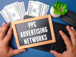 A person holds a small sign that says "PPC Advertising Networks" over a laptop keyboard, with cash, a mouse, and a potted succulent on the desk, highlighting the benefits of using paid ads in digital marketing.